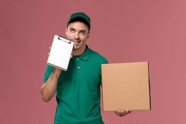 Front view male courier in green uniform holding food box along with little notepad on light-pink background 