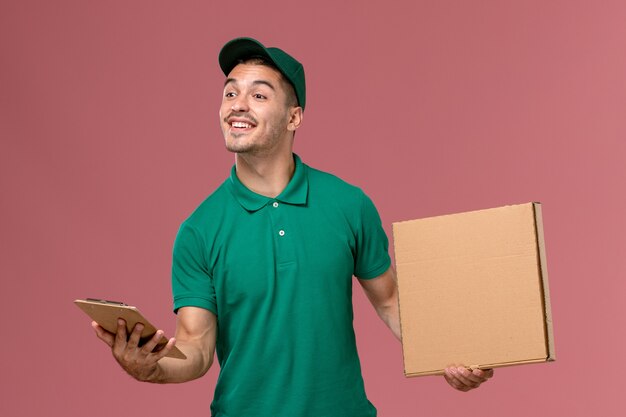 Front view male courier in green uniform holding food box along with notepad on light-pink background 
