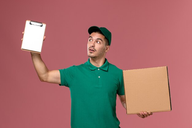 Front view male courier in green uniform holding food box along with notepad on light pink background 