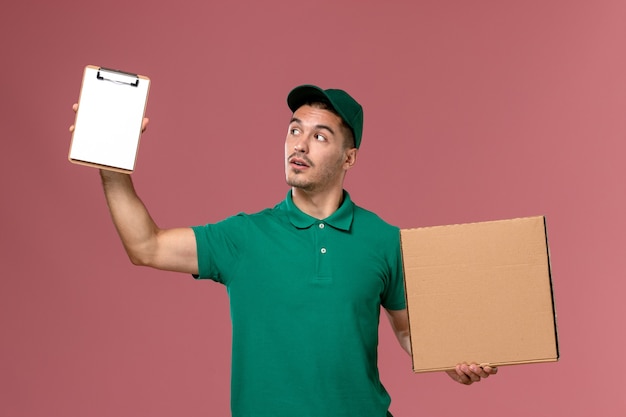 Курьер-мужчина в зеленой форме, держа коробку с едой и блокнотом на светло-розовом фоне, вид спереди