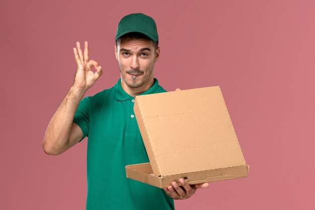 Front view male courier in green uniform holding food box and opening it on light pink background 