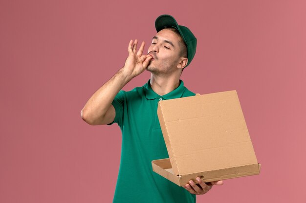 Free photo front view male courier in green uniform holding food box and opening it on light-pink desk