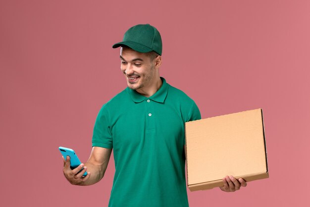 フードボックスを保持し、ピンクの背景に彼の電話を使用して緑の制服を着た男性の宅配便の正面図