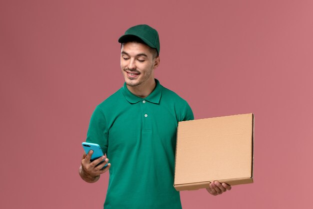 Курьер-мужчина, вид спереди в зеленой форме, держит коробку с едой и разговаривает по телефону на розовом фоне
