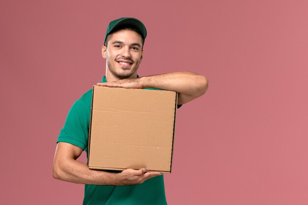 Front view male courier in green uniform holding food box with a smile on the pink background 