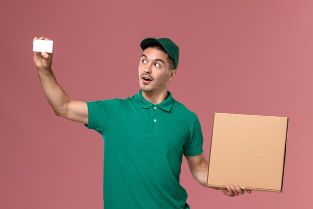 Front view male courier in green uniform holding food box with white card on the pink background 