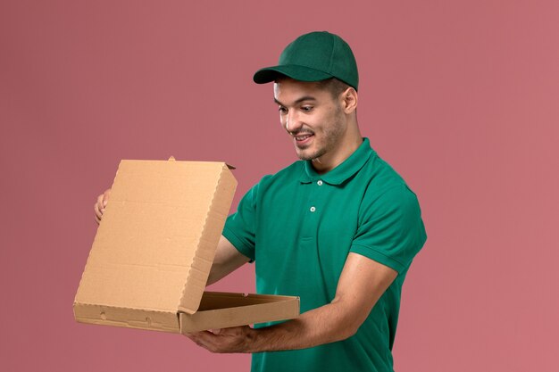 Front view male courier in green uniform holding food delivery box on pink background