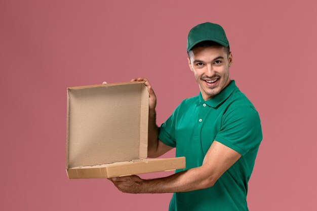 Front view male courier in green uniform holding food delivery box on the pink background