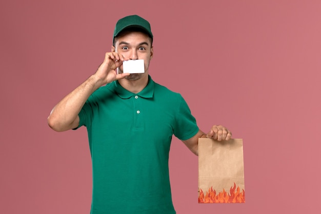 Front view male courier in green uniform holding food package and card on the pink background 