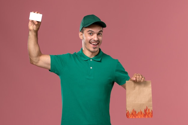ピンクの背景に食品パッケージとカードを保持している緑の制服の正面図男性宅配便