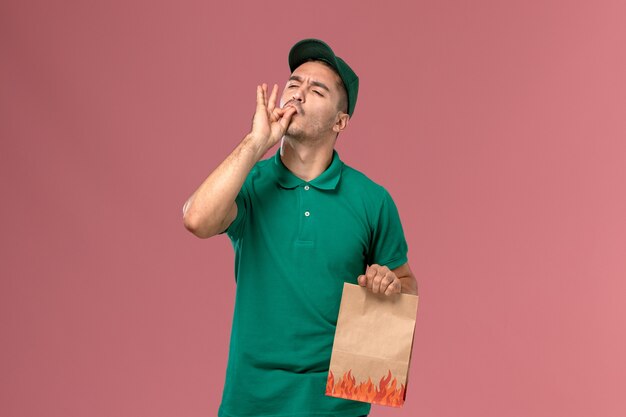 Front view male courier in green uniform holding food package on light pink background   