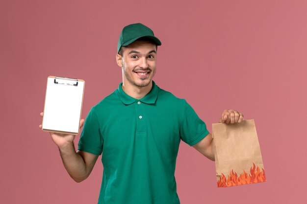 ピンクの背景に食品パッケージとメモ帳を保持している緑の制服の正面図男性宅配便
