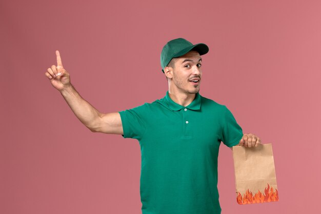 淡いピンクの背景に上げられた指で食品パッケージとposignを保持している緑の制服の正面図男性宅配便