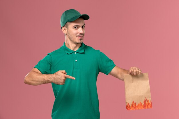 Front view male courier in green uniform holding food package and posing on light-pink background