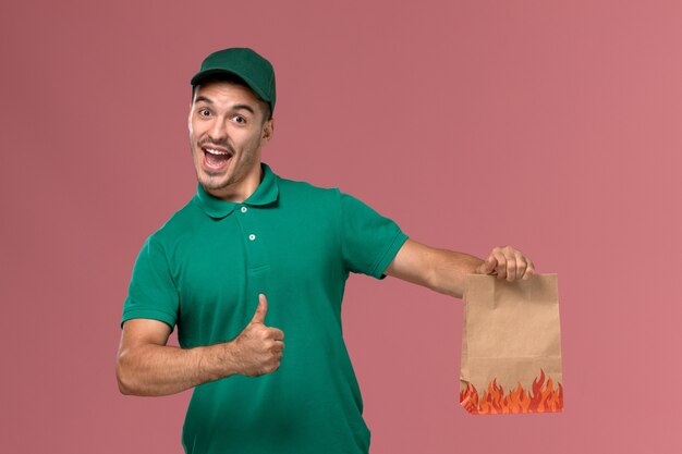 Front view male courier in green uniform holding food package and rejoicing on light-pink background