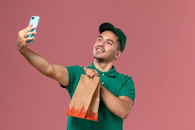 Front view male courier in green uniform holding food package and taking photo with it on the pink background 