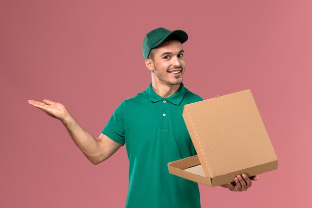 Front view male courier in green uniform holding and opening food box with smile on light pink background   