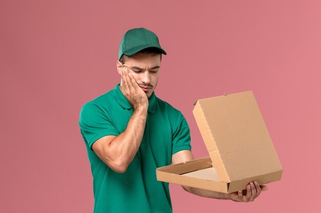 Курьер-мужчина, вид спереди в зеленой форме, держит и открывает коробку с едой с подчеркнутым выражением лица на светло-розовом фоне