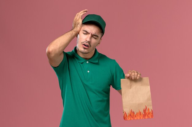 Front view male courier in green uniform holding paper food package having headache on light-pink background   