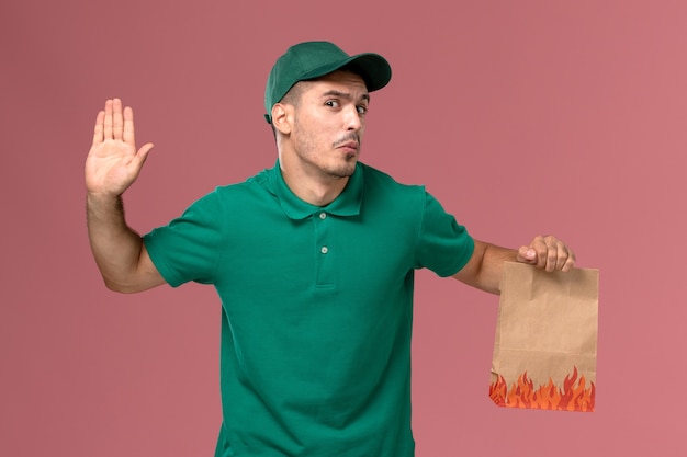 Front view male courier in green uniform holding paper food package on the light pink background   