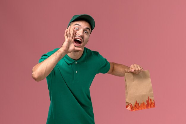 Front view male courier in green uniform holding paper food package screaming on light-pink background   