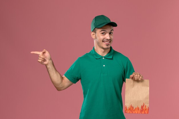 淡いピンクの背景に笑顔で紙の食品パッケージを保持している緑の制服の正面図男性宅配便