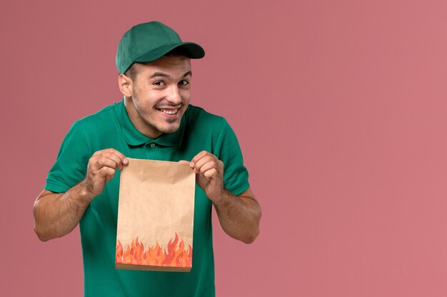 Front view male courier in green uniform holding paper food package with a smile on the pink background   