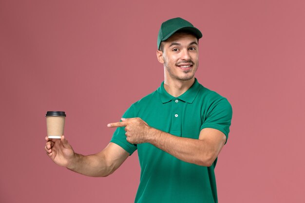 Front view male courier in green uniform smiling and holding  coffee cup on light-pink background  