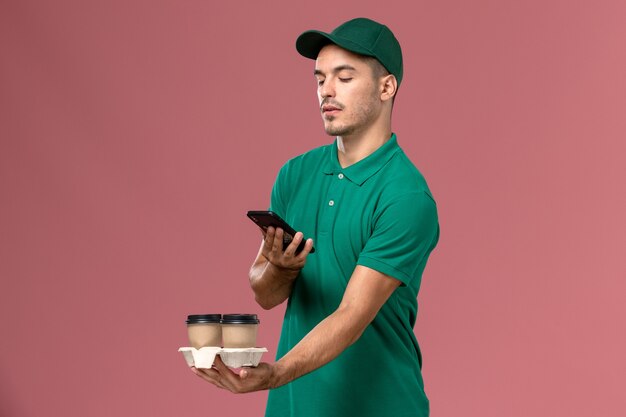 Курьер-мужчина в зеленой форме, фотографирующий кофе на розовом фоне, вид спереди
