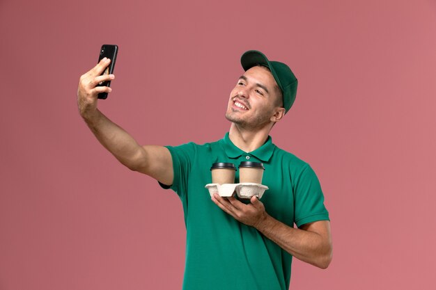 Курьер-мужчина в зеленой форме, фотографирующийся с кофе на розовом фоне, вид спереди