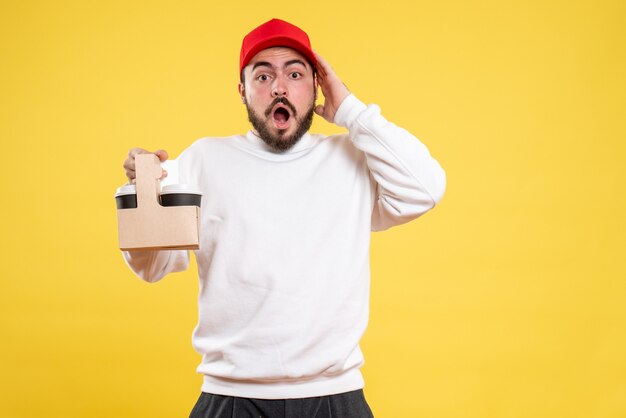 Front view of male courier holding delivery coffee on yellow wall