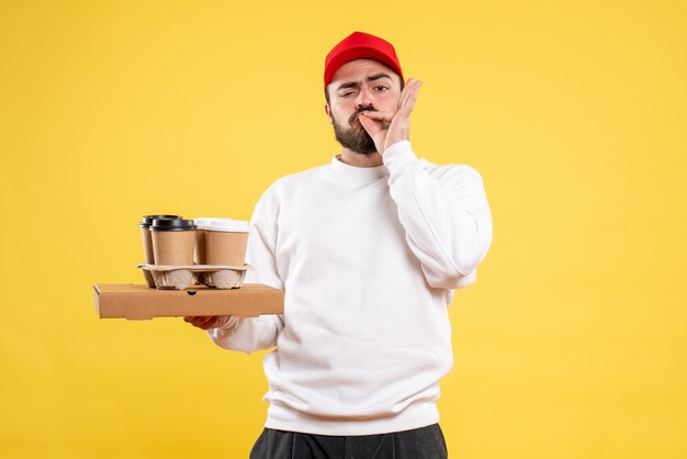 Front view of male courier holding delivery food and coffee on yellow wall