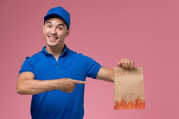 ピンクの壁に紙の食品パッケージを保持している青い制服の正面図男性宅配便、均一なサービスの仕事の配達