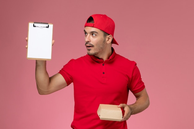 Бесплатное фото Курьер-мужчина вида спереди в красной форме и накидке, держащий маленький пакет доставки с блокнотом на розовом фоне.