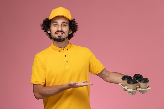 Бесплатное фото Курьер-мужчина в желтой форме, держащий коричневые кофейные чашки на розовой стене, вид спереди