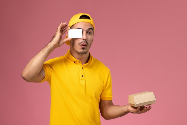 Курьер мужского пола вид спереди в желтой форме держа карточку и небольшой пакет еды на розовом фоне.