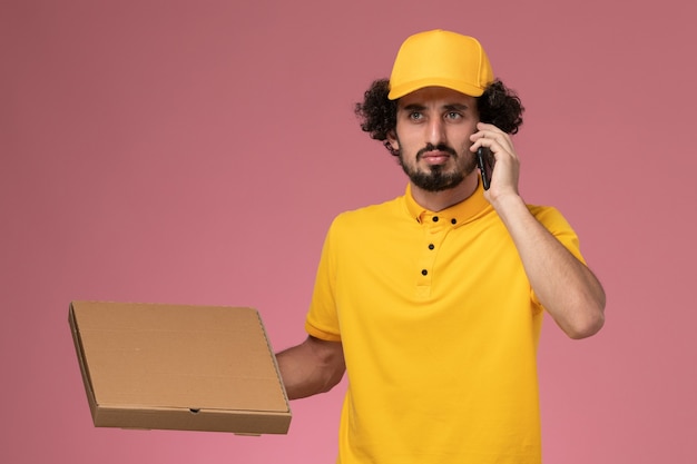 음식 배달 상자를 들고 밝은 분홍색 벽에 전화로 이야기하는 노란색 제복을 입은 전면보기 남성 택배