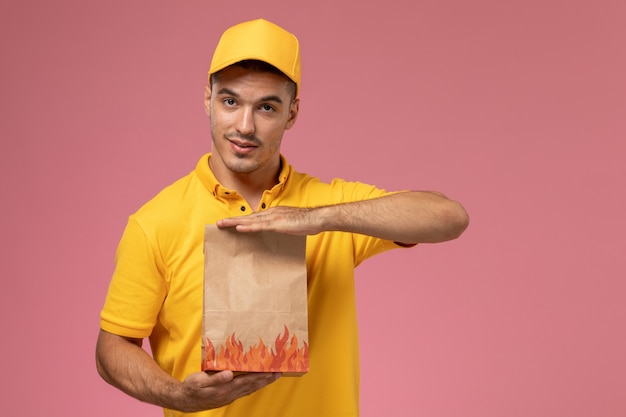 Курьер-мужчина в желтой форме, держа пакет с едой на розовом фоне, вид спереди