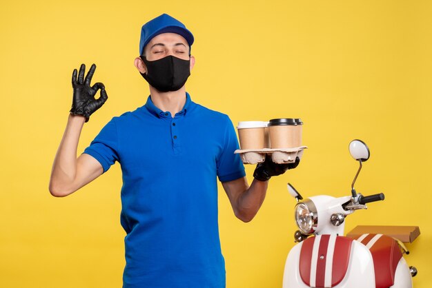 Front view male courier in mask holding coffee on yellow job covid- pandemic delivery uniform work