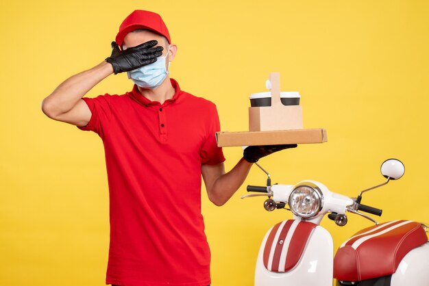 Front view male courier in mask with delivery coffee and box covering his eyes on yellow color service covid- virus job uniform pandemic work