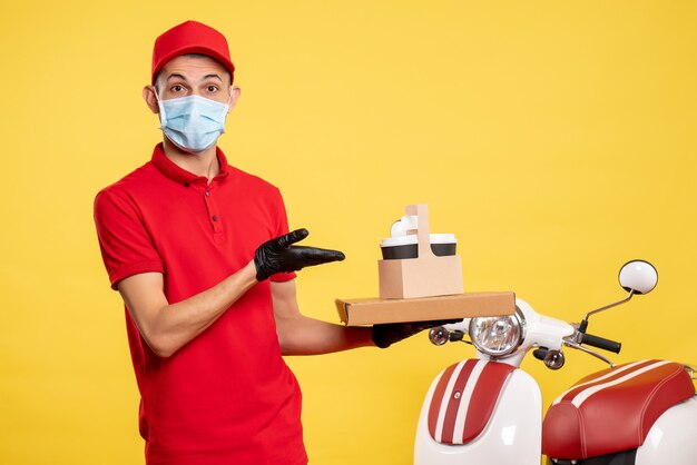 Free photo front view male courier in mask with delivery coffee and box on a yellow color service covid- virus job uniform pandemic work