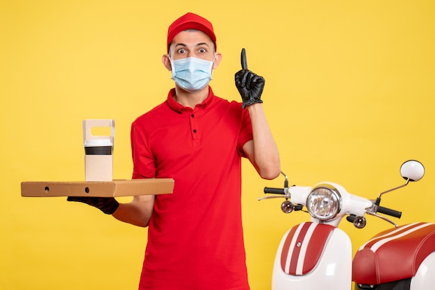 Front view male courier in mask with delivery coffee and box on yellow job color service covid- virus pandemic work uniform