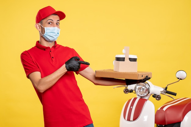 Front view male courier in mask with delivery coffee and box on a yellow service covid pandemic color virus job work uniform