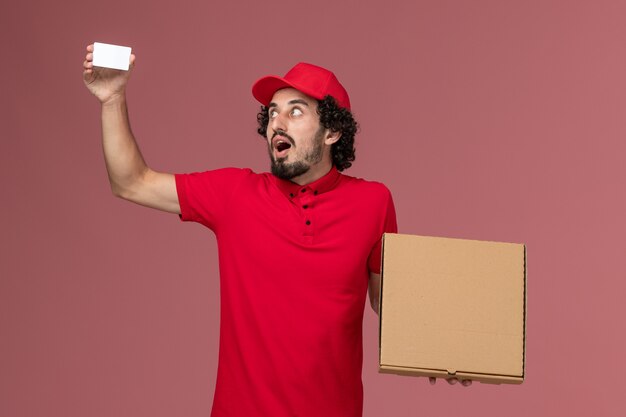 Front view male courier in red shirt and cape holding delivery food box and card on pink wall