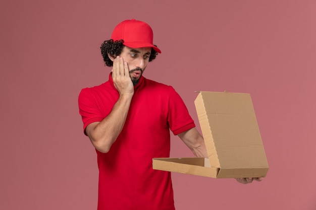 Курьер-мужчина в красной рубашке и плаще, вид спереди, держит пустую коробку для еды на розовой стене
