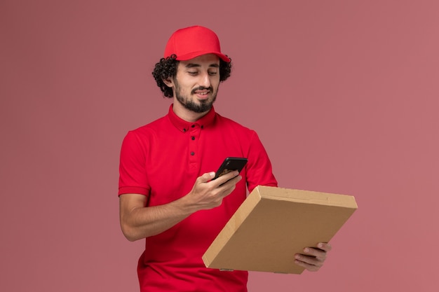 Front view male courier in red shirt and cape holding empty delivery food box taking picture of it on pink wall