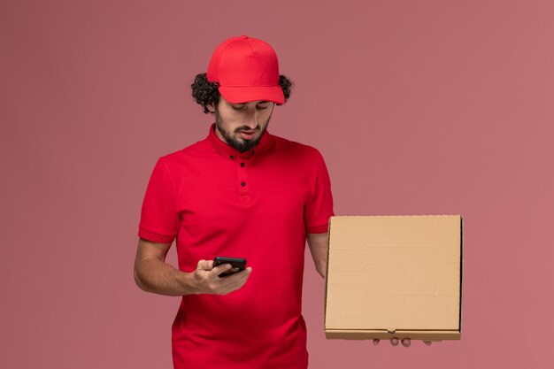 Front view male courier in red shirt and cape holding empty delivery food box with phone on pink wall