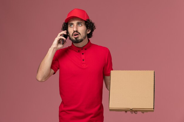 Курьер-мужчина в красной рубашке и плаще, держащий пустую коробку для доставки еды, разговаривает по телефону на розовой стене
