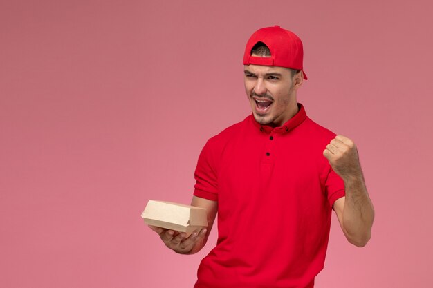 Front view of male courier in red uniform and cap holding little delivery package cheering on the pink wall