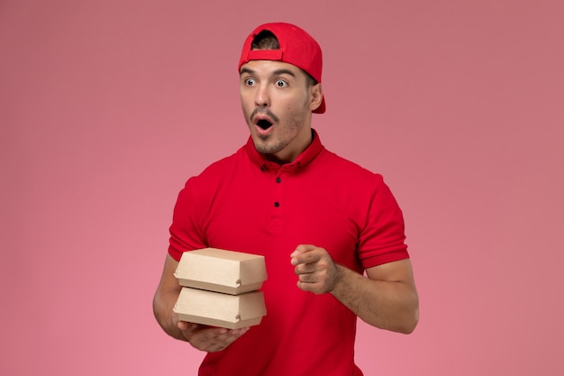 Front view of male courier in red uniform and cap holding little delivery package on light pink wall